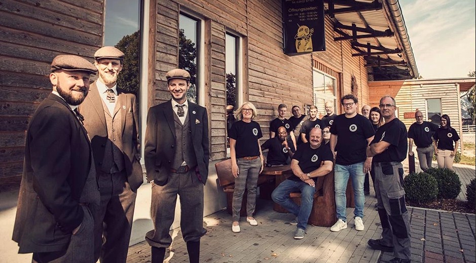 Gruppe von Menschen in schwarzen T-Shirts und formeller Kleidung vor einem Holzgebäude, die lächeln und zusammen posieren.