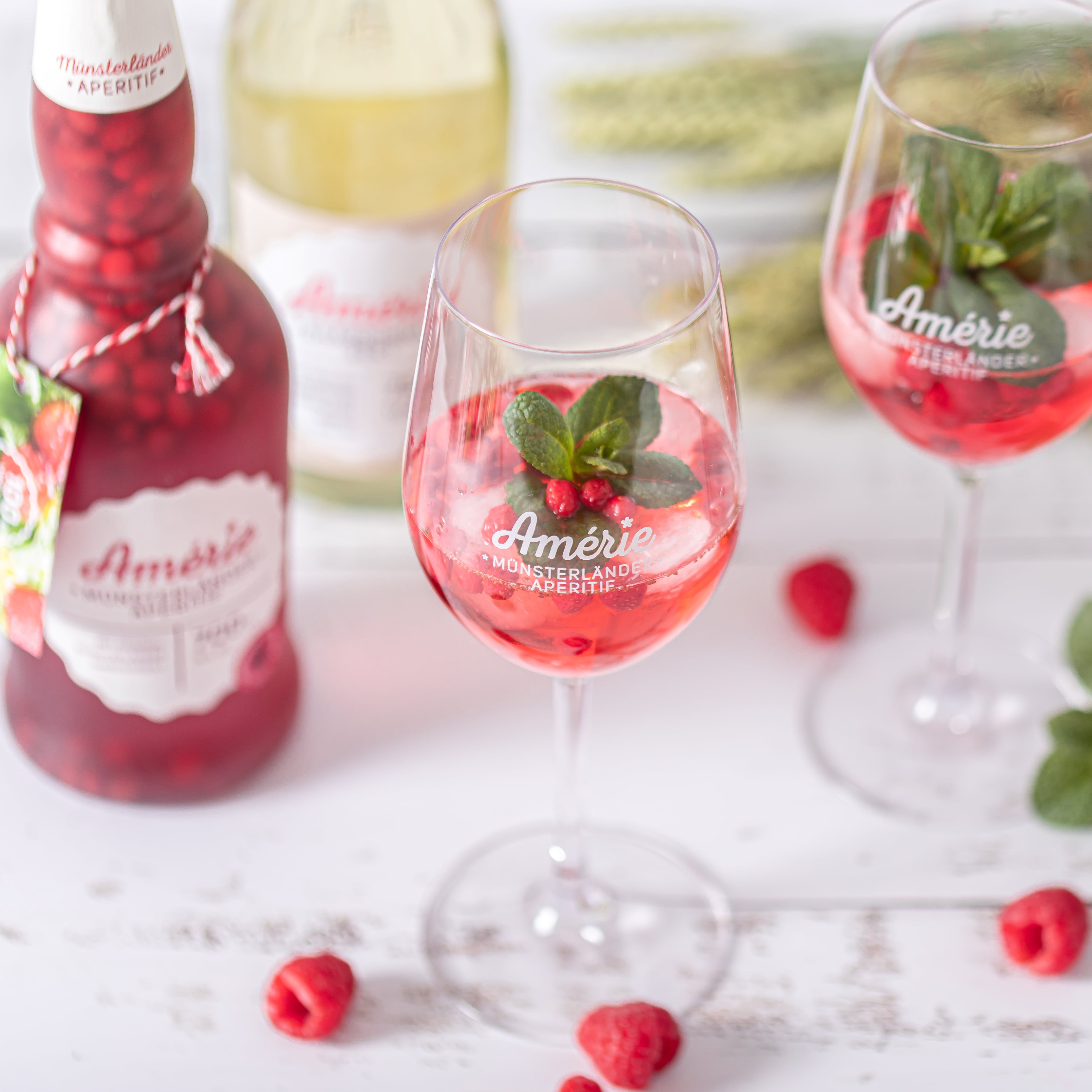 Amérie Münsterländer Aperitif in Markengläsern mit frischen Himbeeren und Minze auf einem rustikalen weißen Holztisch.